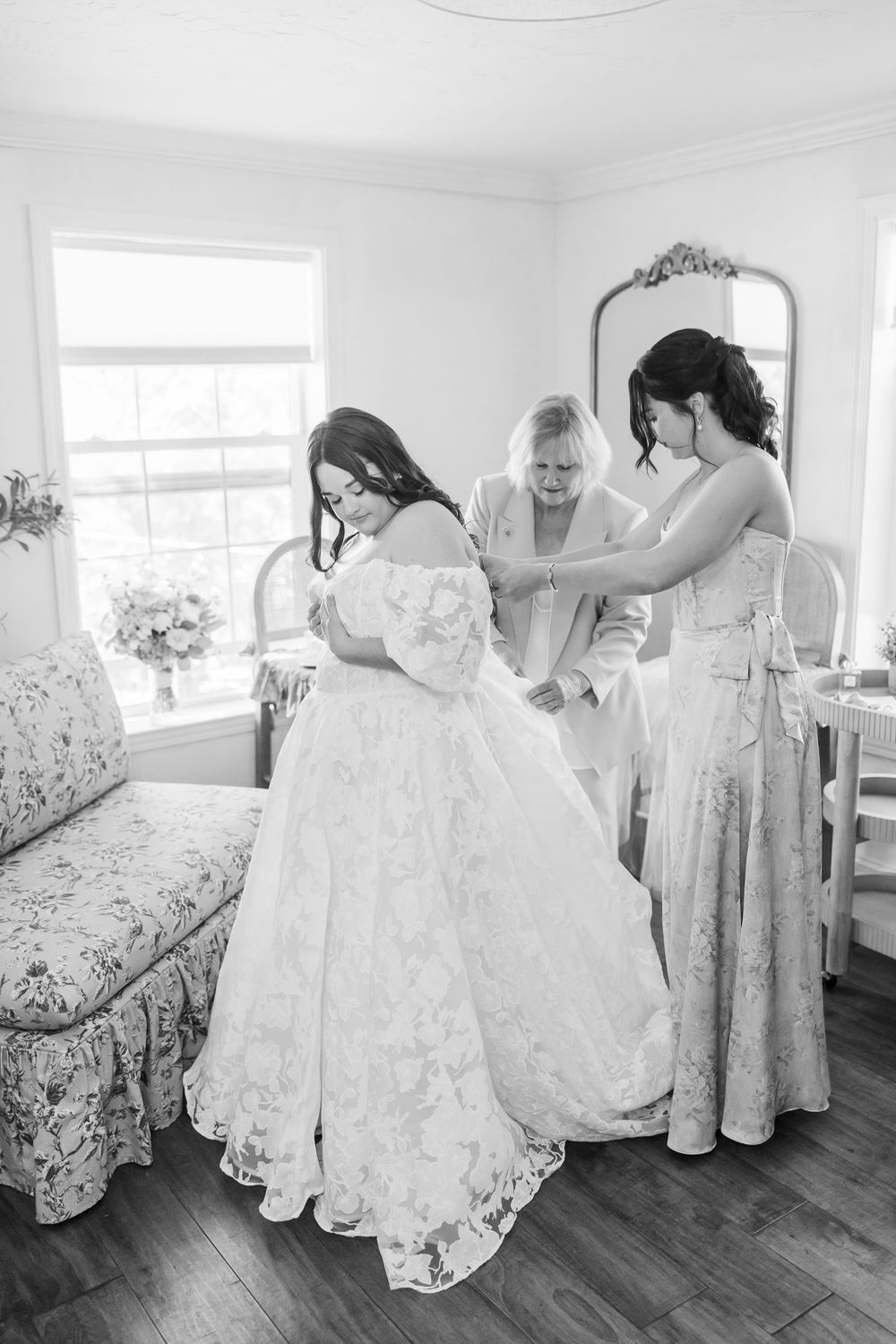 Black and white photo of bride in lace ball gown being helped by two women in bright bridal suite.