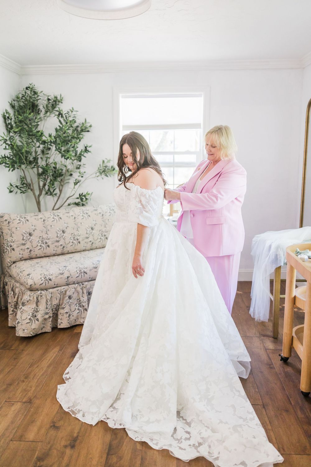 A woman in a pink suit helps a bride in a lace off-shoulder wedding gown get dressed in a bright white room.
