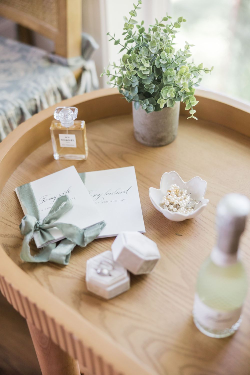 Wooden tray with wedding rings, perfume, bridesmaid cards tied with sage ribbon, and small plant.