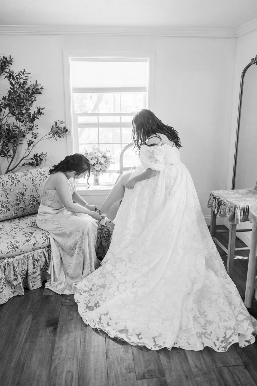 Black and white photo of bridesmaid helping bride with shoes while seated near bright window.