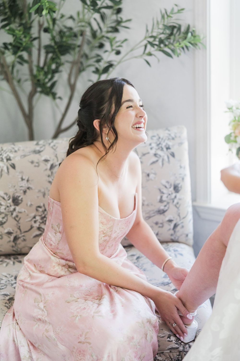 Laughing bridesmaid in pink floral dress sits on floral sofa helping with shoes in bright elegant bridal suite.