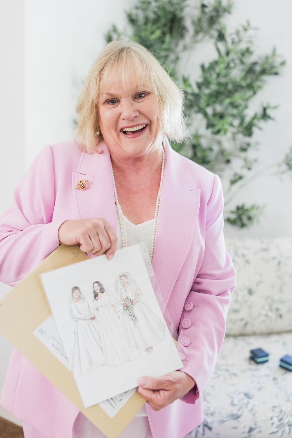 Smiling woman in pink blazer proudly holds pencil sketch drawings of a wedding party portrait.