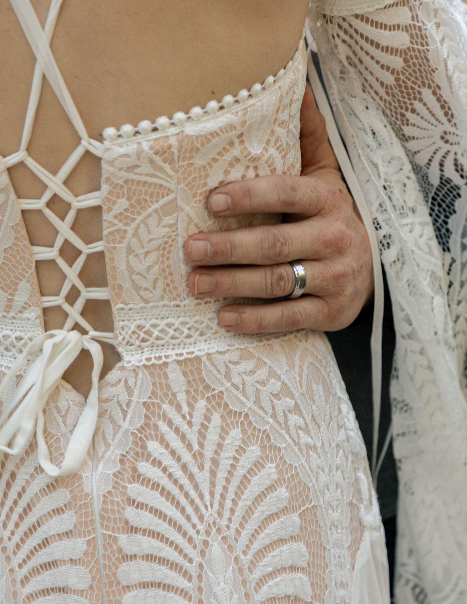 Close-up of a hand with a silver wedding band resting on an intricate white lace bridal gown back.