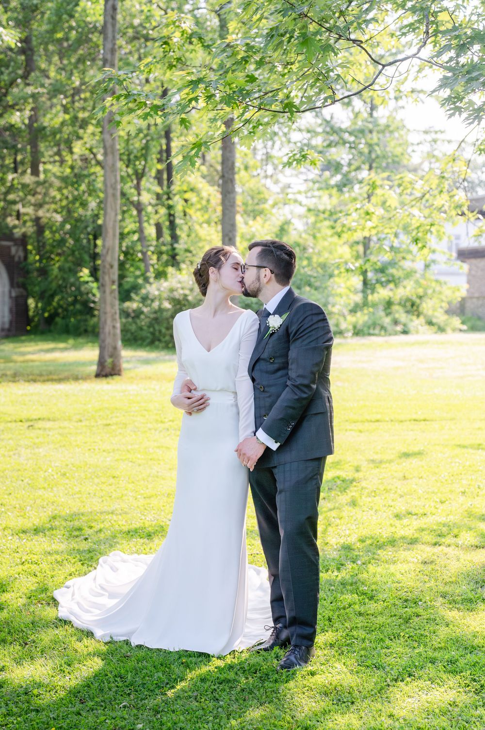 Bride and groom share a kiss in a lush green garden, she in a white gown, he in a dark suit with boutonniere.