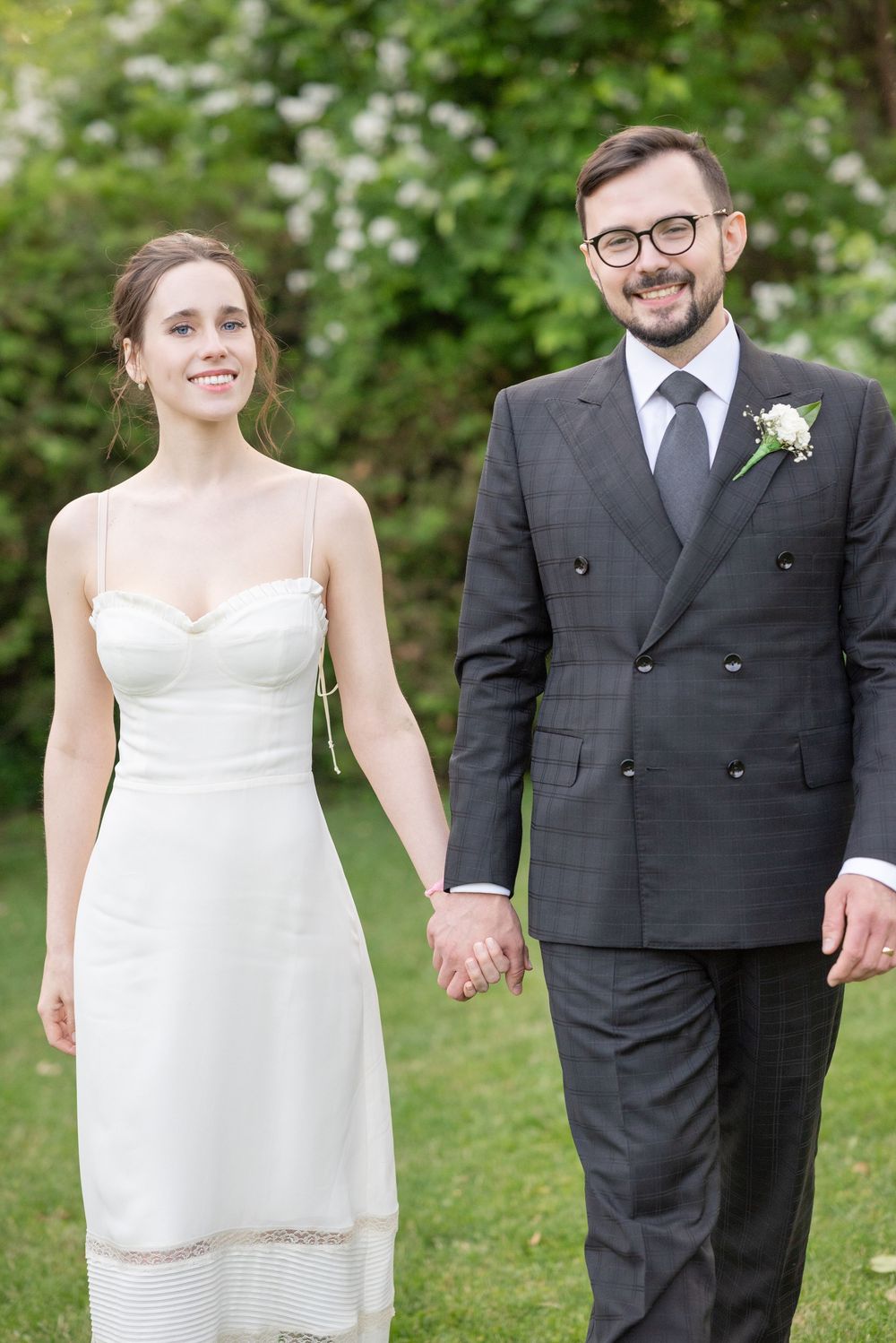 Smiling bride in white dress holds hands with groom in dark suit and glasses outdoors on wedding day.