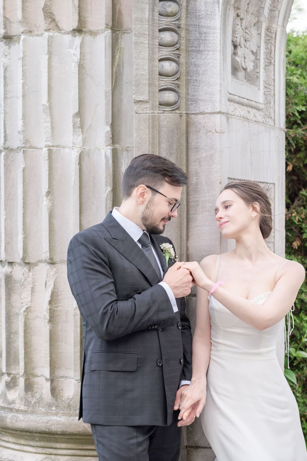 Bride and groom sharing a tender moment by stone columns, she in a white slip dress, he in a gray suit with boutonniere.