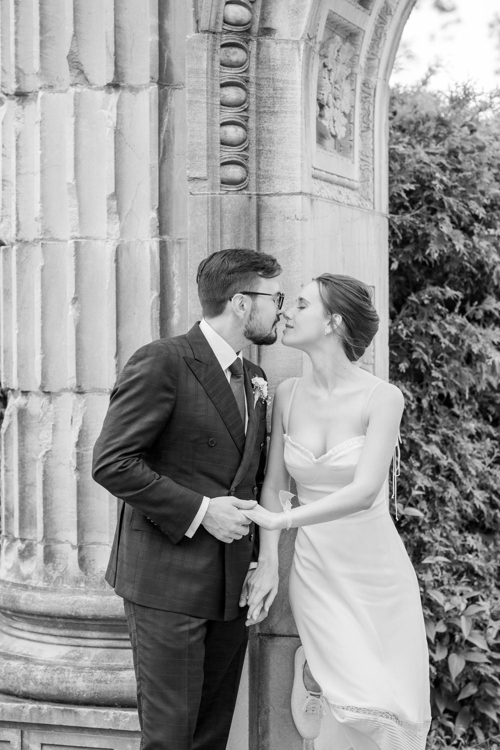 Black and white photo of a bride and groom sharing a romantic moment beside ornate stone columns at their wedding.