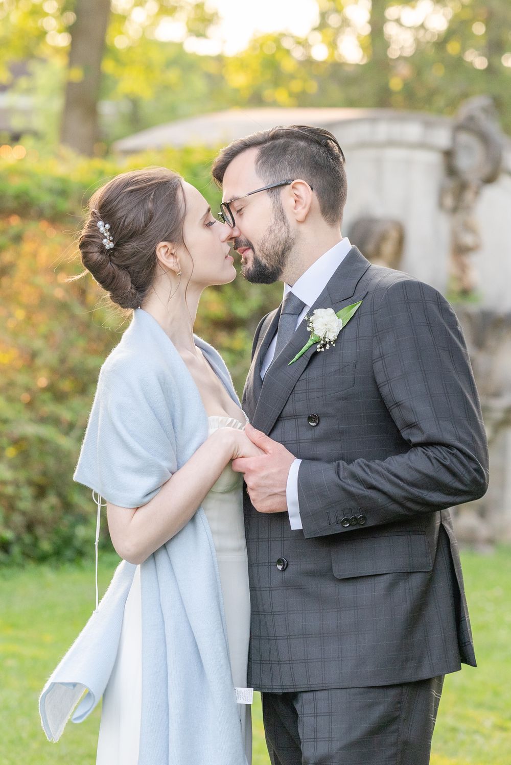 A bride and groom share a romantic kiss outdoors, dressed in wedding attire with lush greenery in the background.