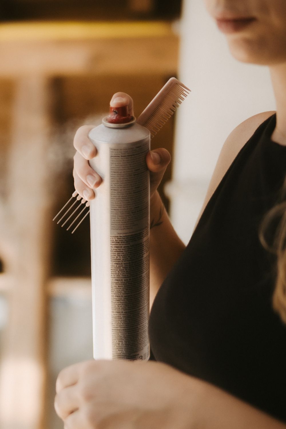 Hairstylist holding a hair spray can and comb in a salon setting, wearing a black apron.