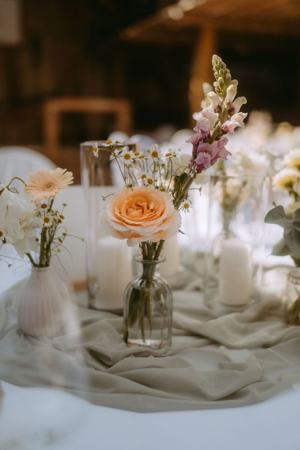 Elegant wedding table with peach roses, daisies, snapdragons in glass vases, white candles on sage green linen.