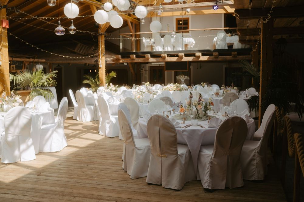 Elegant wedding reception venue with white chair covers, round tables, and hanging paper lantern decorations on wooden deck.