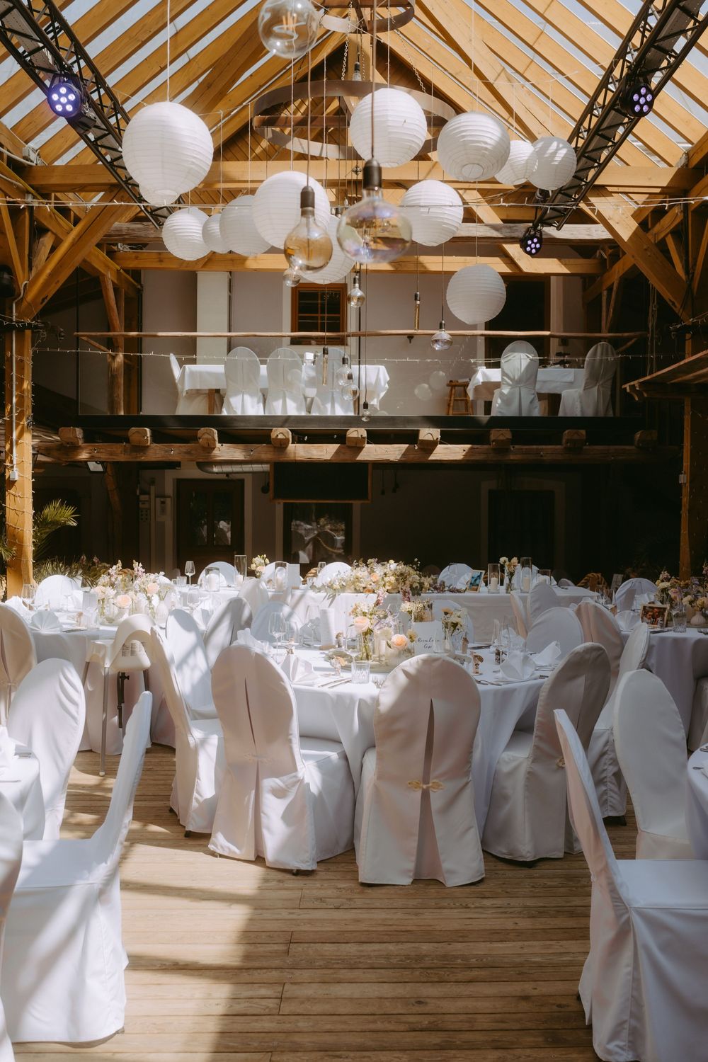 Elegant barn wedding reception with white chair covers, round tables, and white paper lantern ceiling decorations.