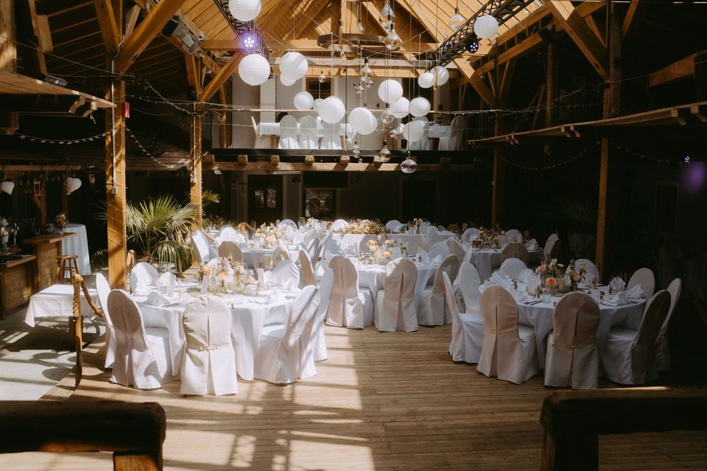 Rustic barn wedding reception with round tables, white chair covers, and globe lanterns hanging from wooden beams.