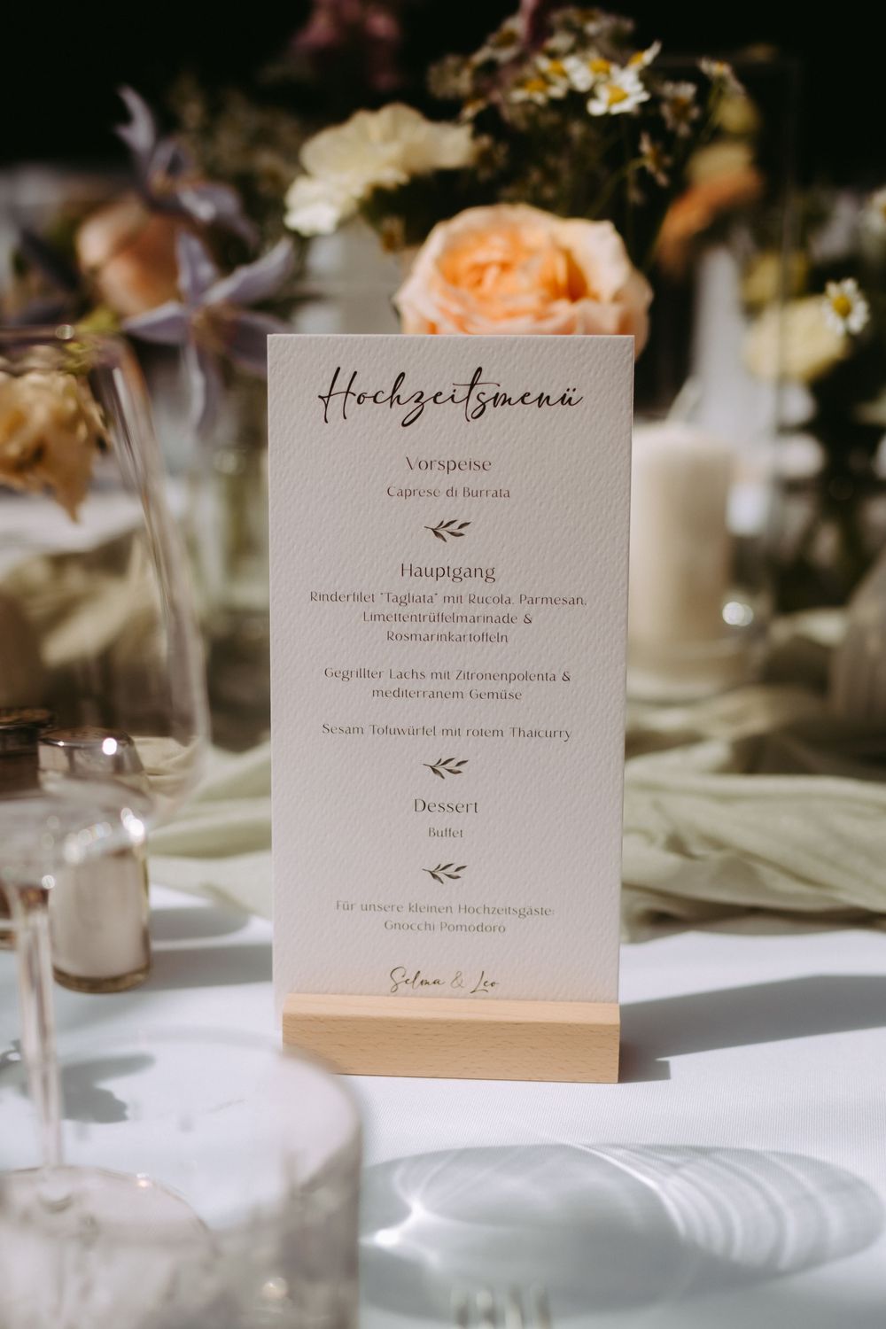 Elegant wedding menu card in German displayed on a wooden stand surrounded by floral arrangements at a reception table.