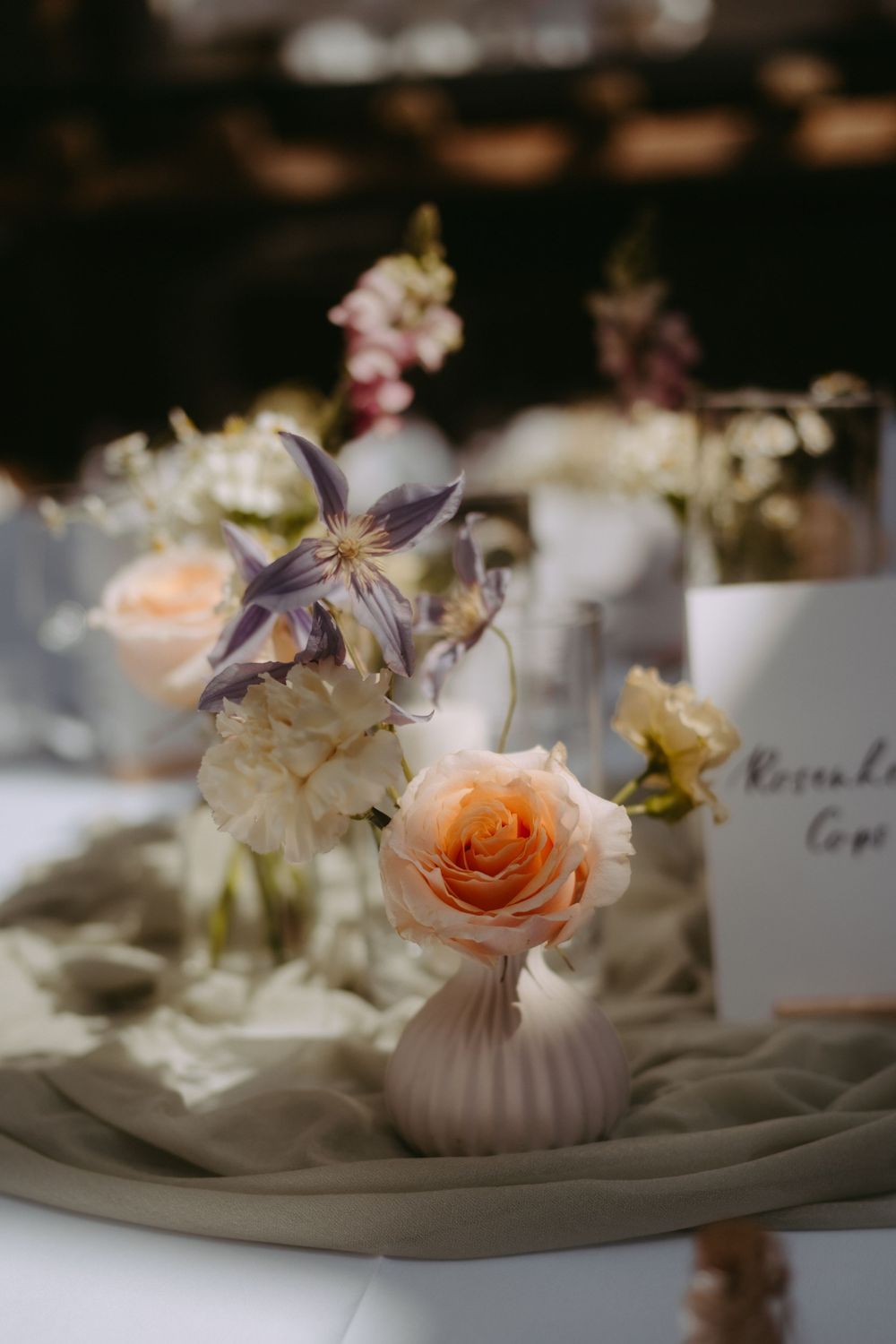 Elegant wedding table centerpiece with peach roses and purple clematis in a white ribbed vase on sage green linen.
