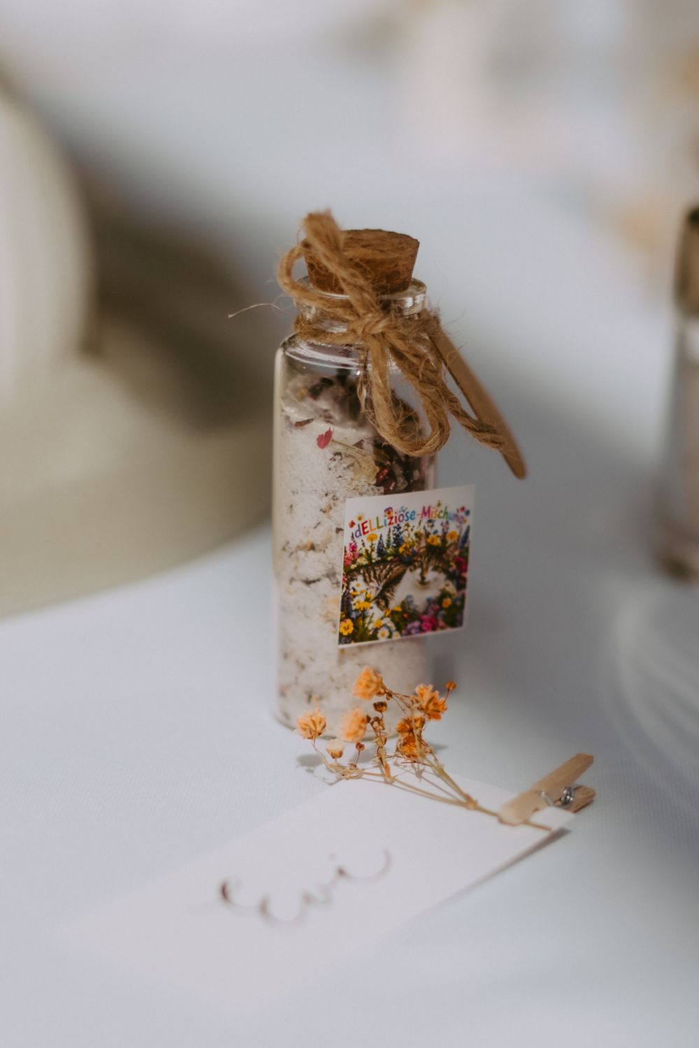 Small glass bottle with cork stopper, twine bow, floral label, and dried wildflowers, displayed on white surface.