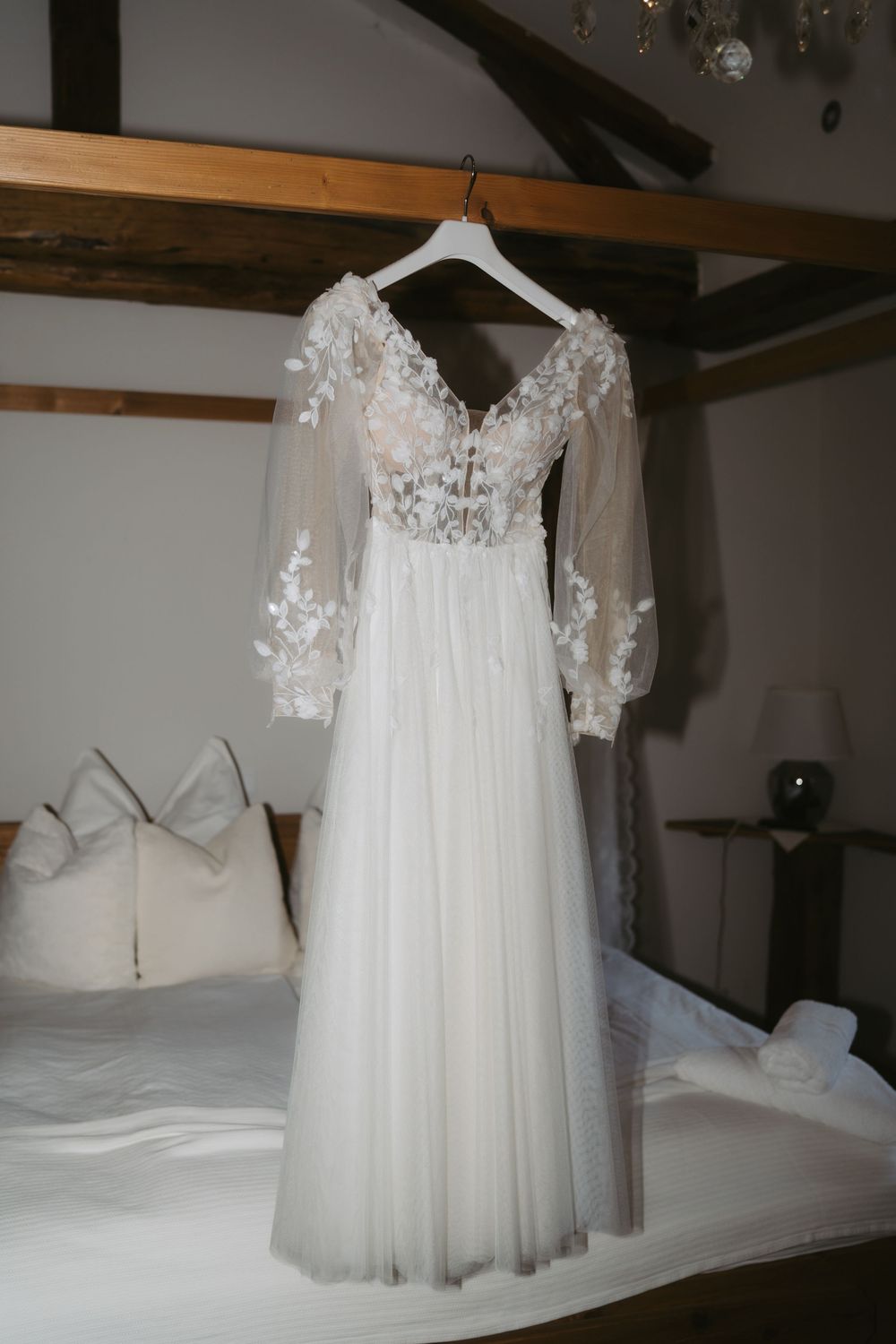 Romantic floral lace wedding dress with sheer long sleeves hanging on a hanger above a white bed.