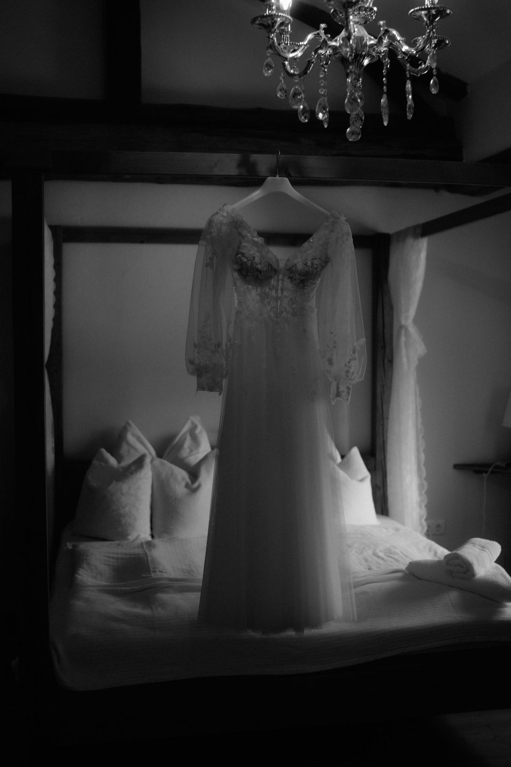 Black and white photo of a lace wedding dress hanging on a four-poster bed beneath a crystal chandelier.