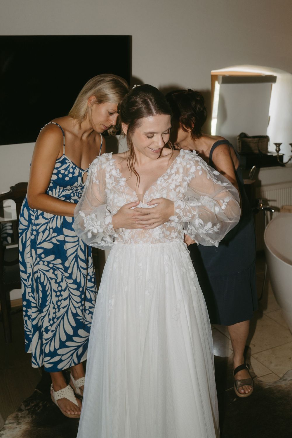 Bride in a lace floral wedding gown being helped by two women in a bridal preparation room.