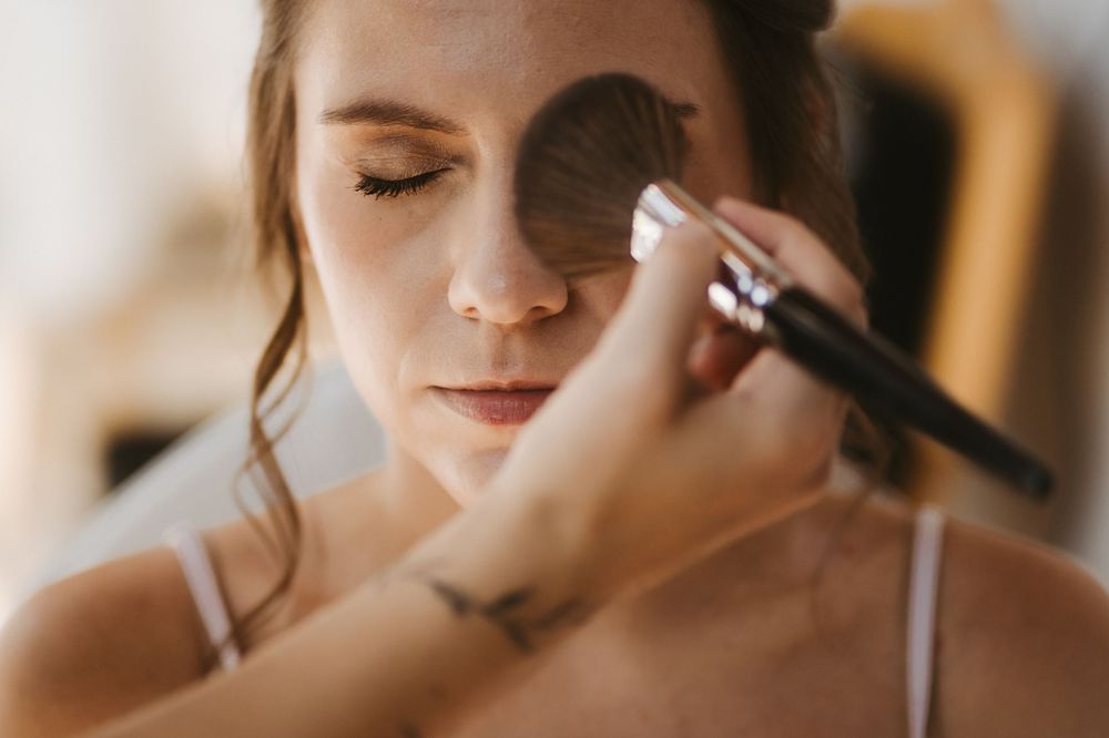 A makeup artist applies powder to a bride's face with a large brush during bridal beauty preparation.