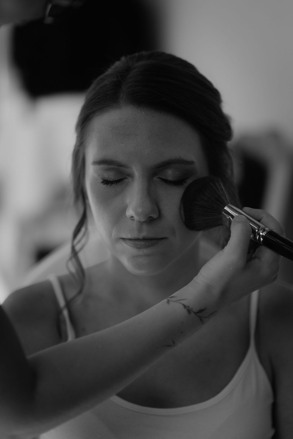 Black and white photo of a bride having makeup applied with a brush before her wedding ceremony.