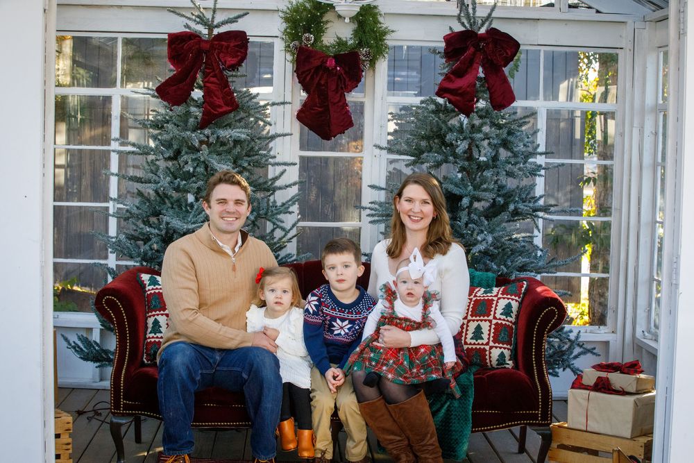 Family of four posing in front of Christmas trees with red bows on a festive holiday porch setting.