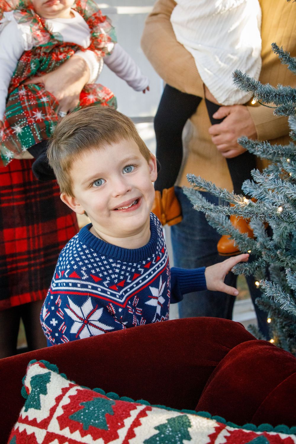 A smiling boy in a Nordic sweater touches a Christmas tree, surrounded by family in festive attire.