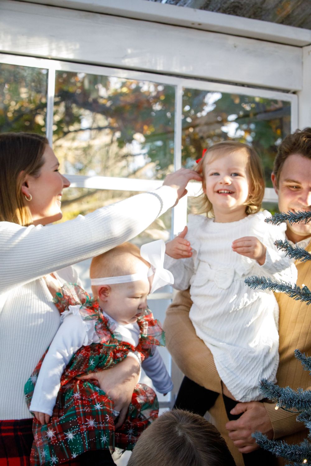 Happy family with young children celebrating Christmas outdoors, holding a festive wreath near a sunlit window.