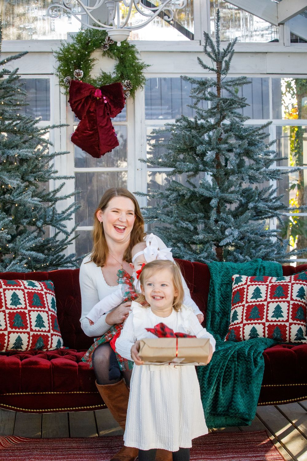 Mother and daughter in white dresses celebrate Christmas with gifts, red sofa, wreath, and festive trees in a decorated room.