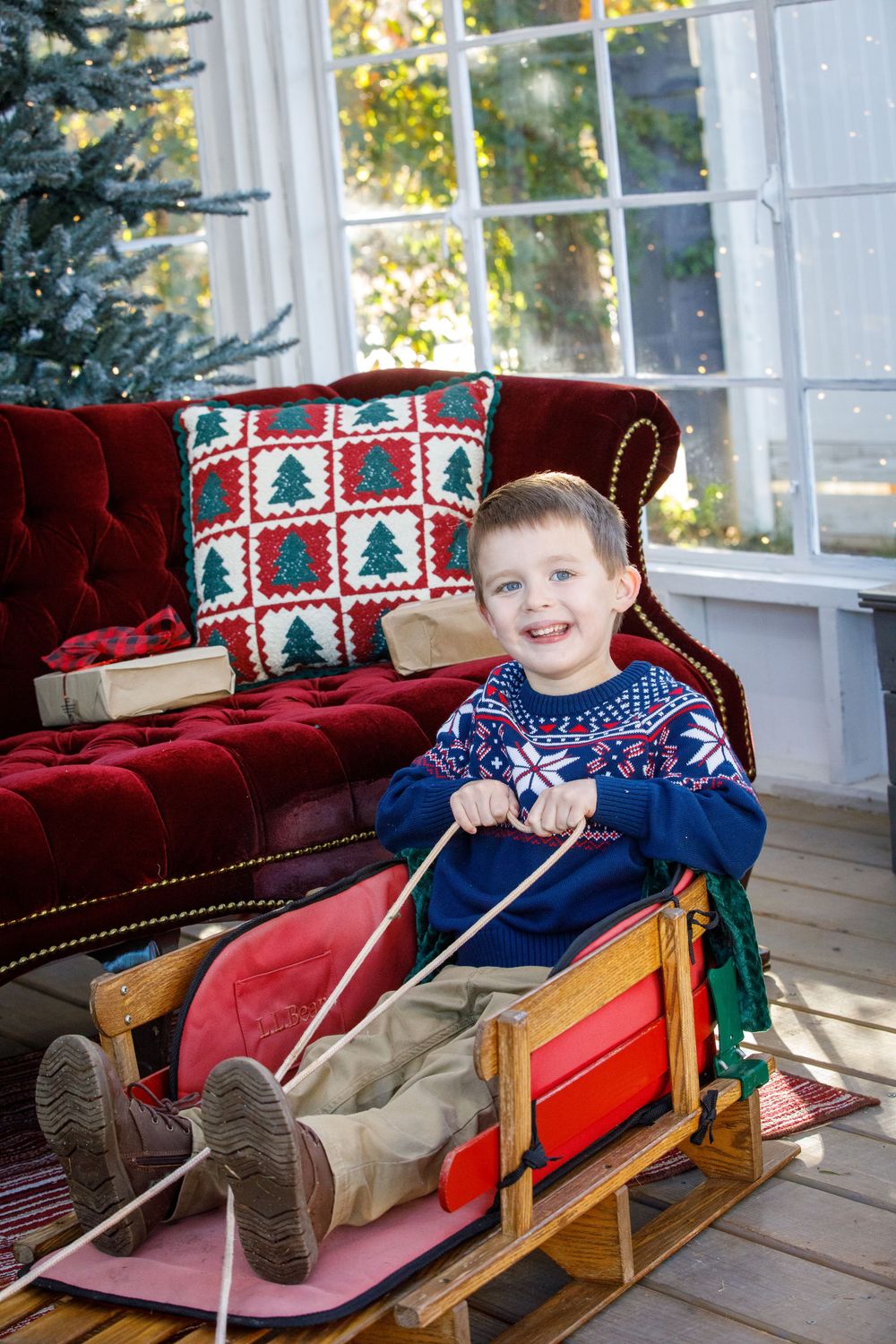 Smiling boy in holiday sweater sitting on wooden sled near red velvet sofa with Christmas tree and gifts.