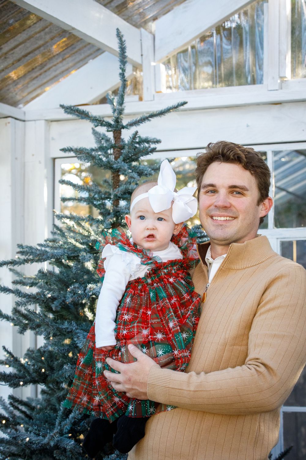 Father holding baby girl in plaid Christmas dress with white bow near decorated outdoor Christmas tree.