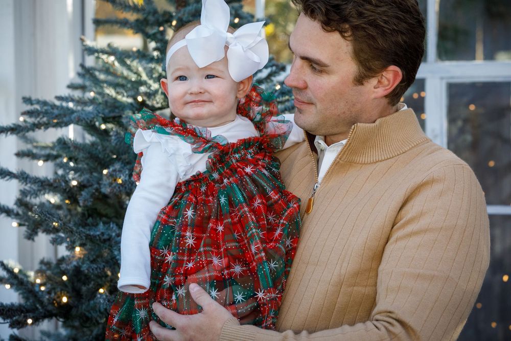 Man holding baby girl in red plaid Christmas dress with white bow near decorated holiday tree.