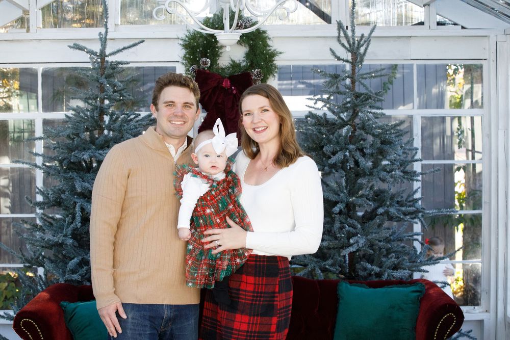 Family holiday portrait with parents holding baby in Christmas dress, posing before decorated trees.