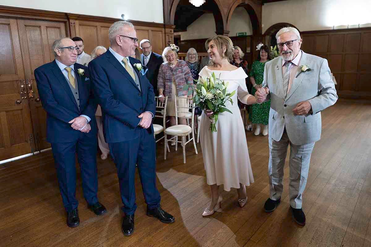 Elegant wedding ceremony with bride holding bouquet, groom, and guests in a wood-paneled hall.