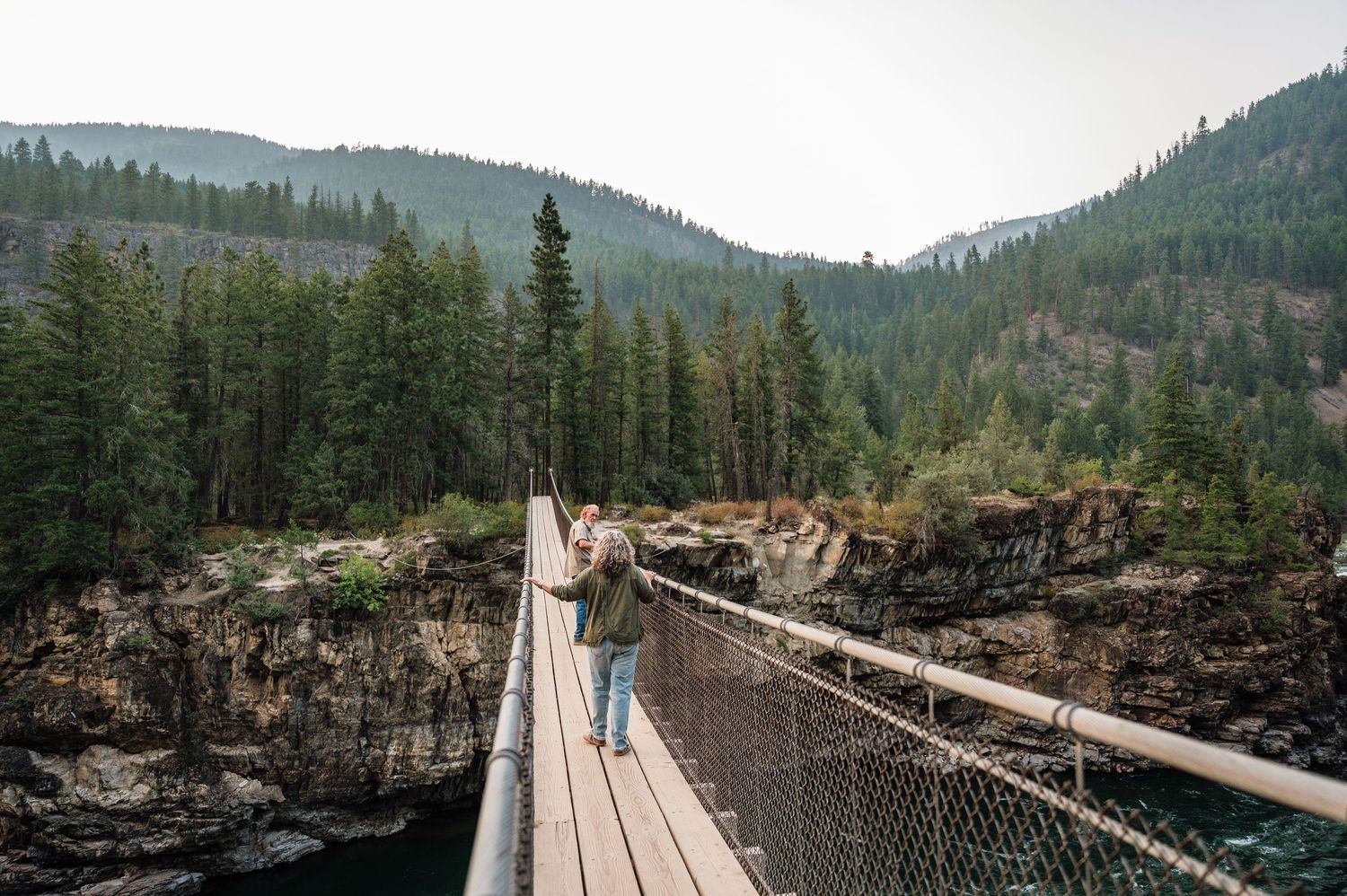 Hiker crosses a wooden suspension bridge over a rocky river gorge surrounded by dense evergreen forest and misty mountains.
