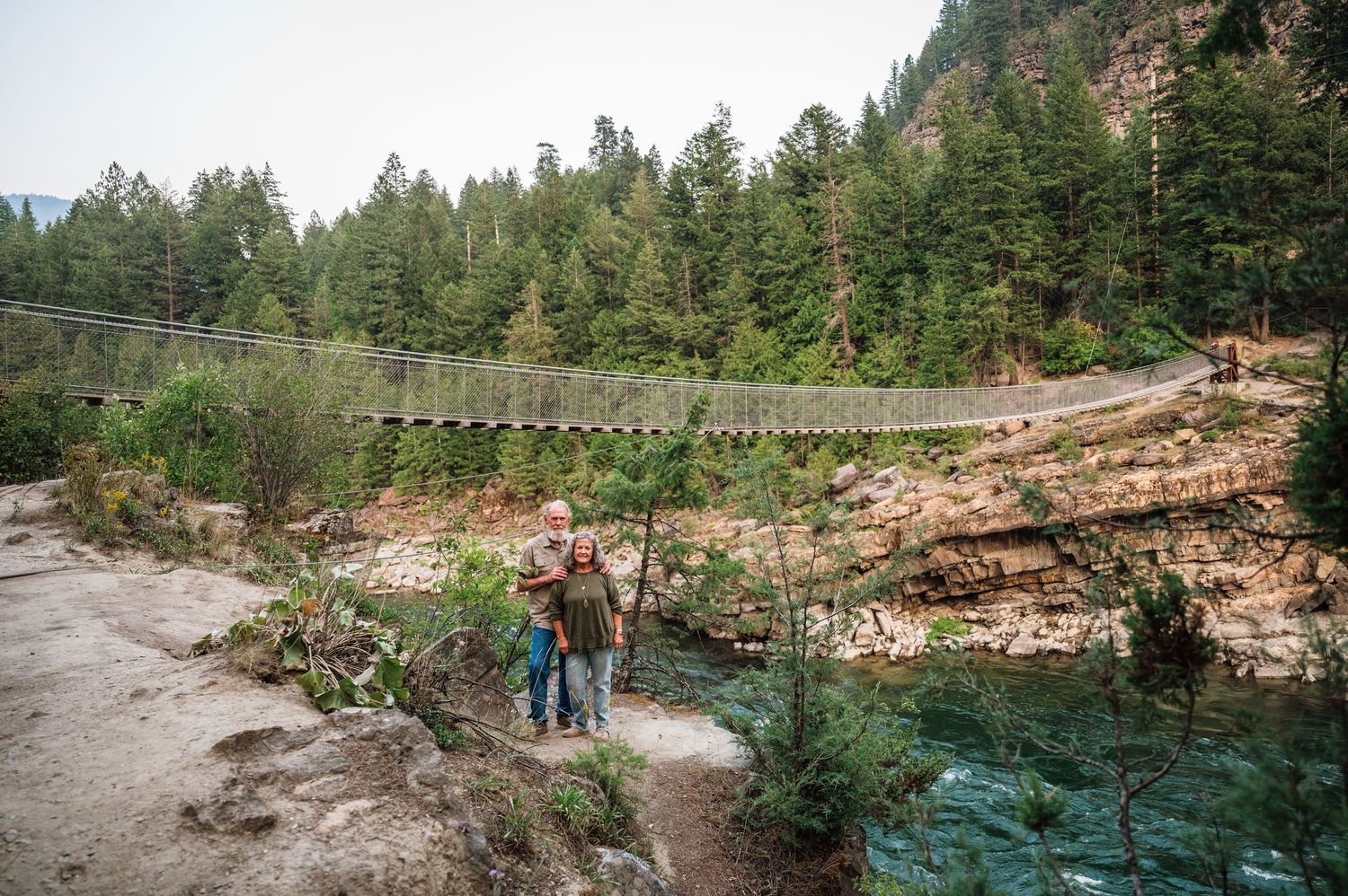 Two hikers stand near a suspension bridge over a turquoise river in a forested canyon.
