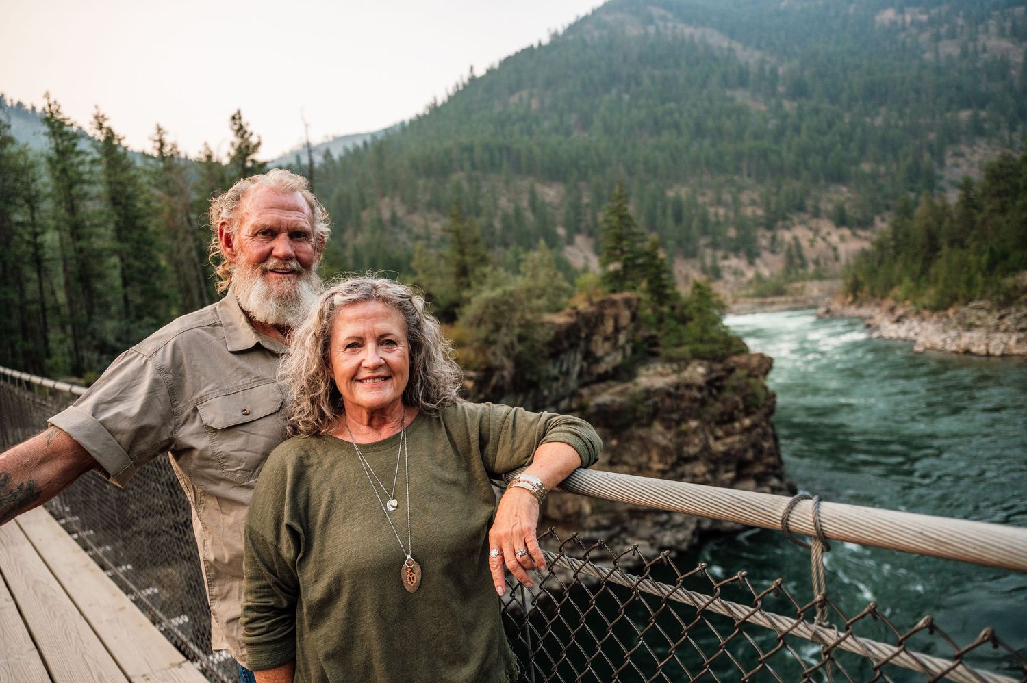 Smiling couple posing on a bridge overlooking a rushing river surrounded by forested mountains.