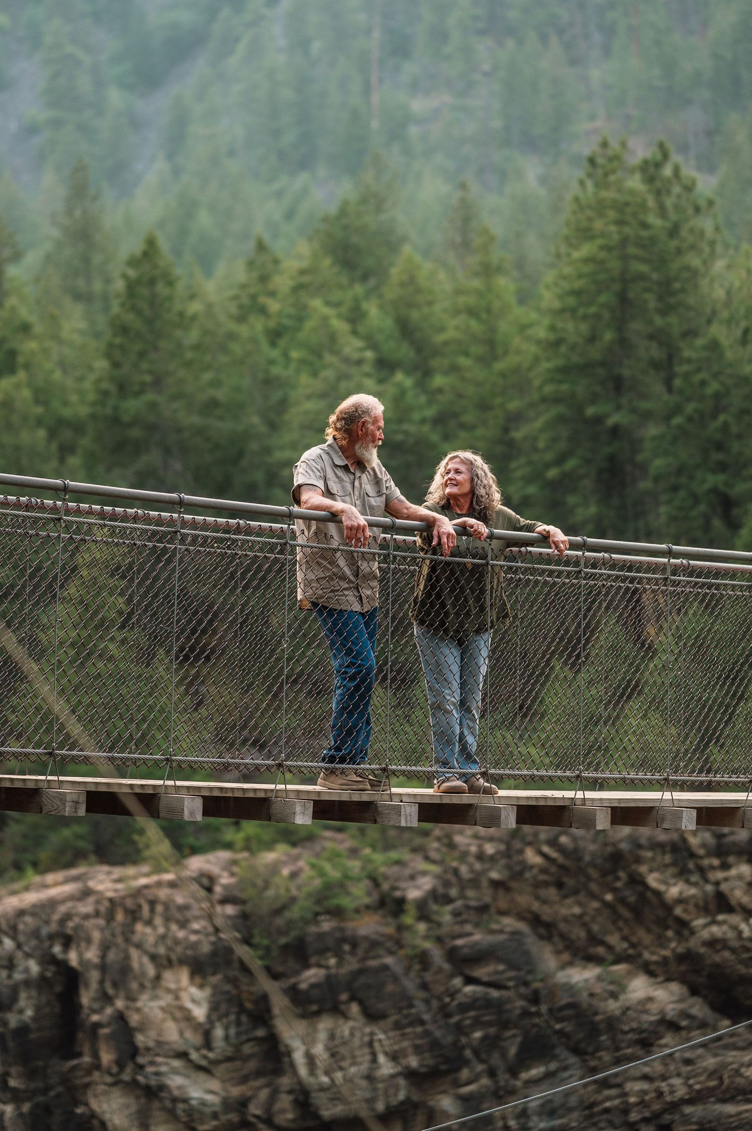 Elderly couple smiling together on a suspension bridge surrounded by lush green forest.