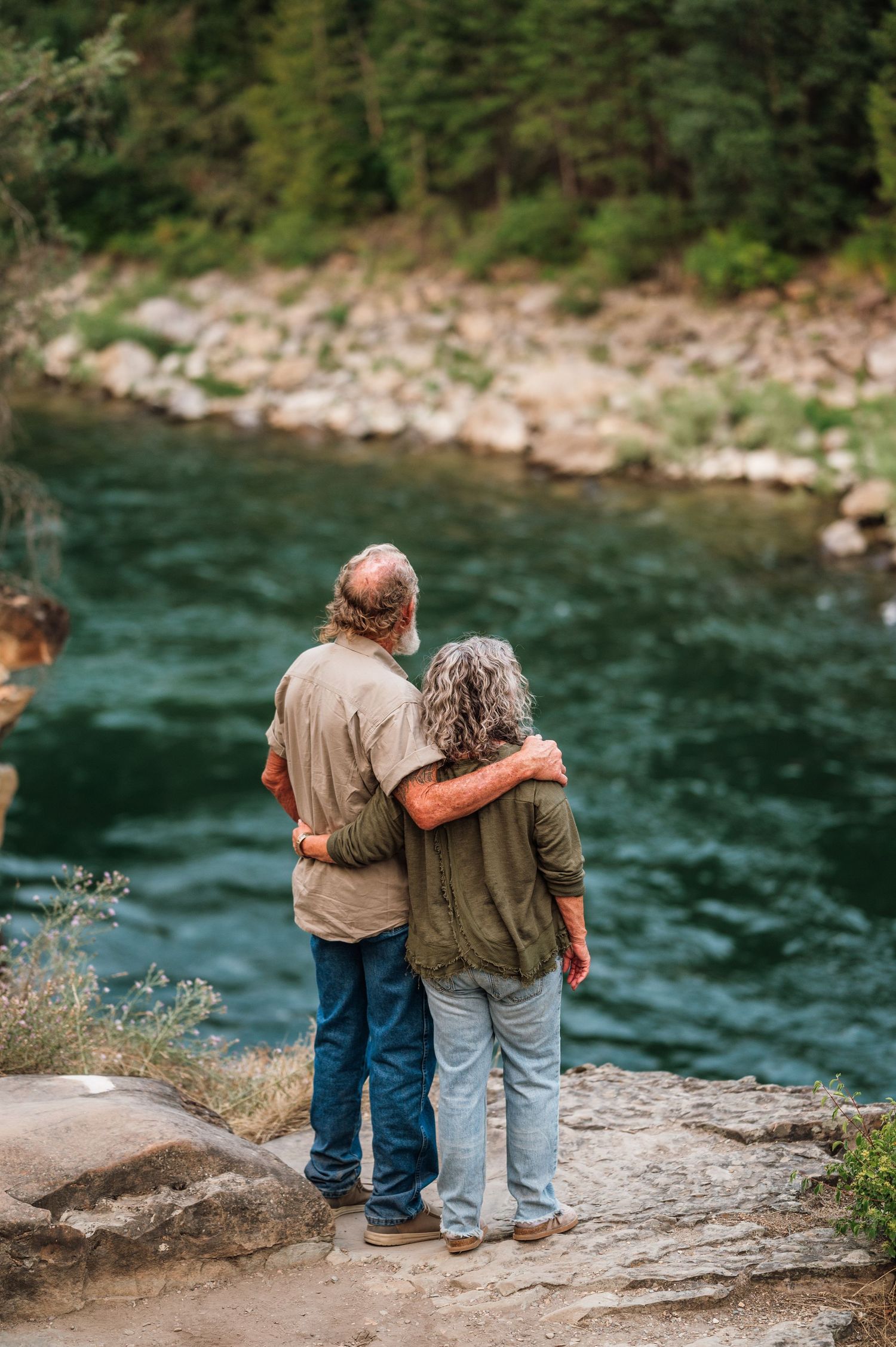 An elderly couple stands embracing on rocky riverbank, gazing at a serene turquoise river surrounded by lush forest.