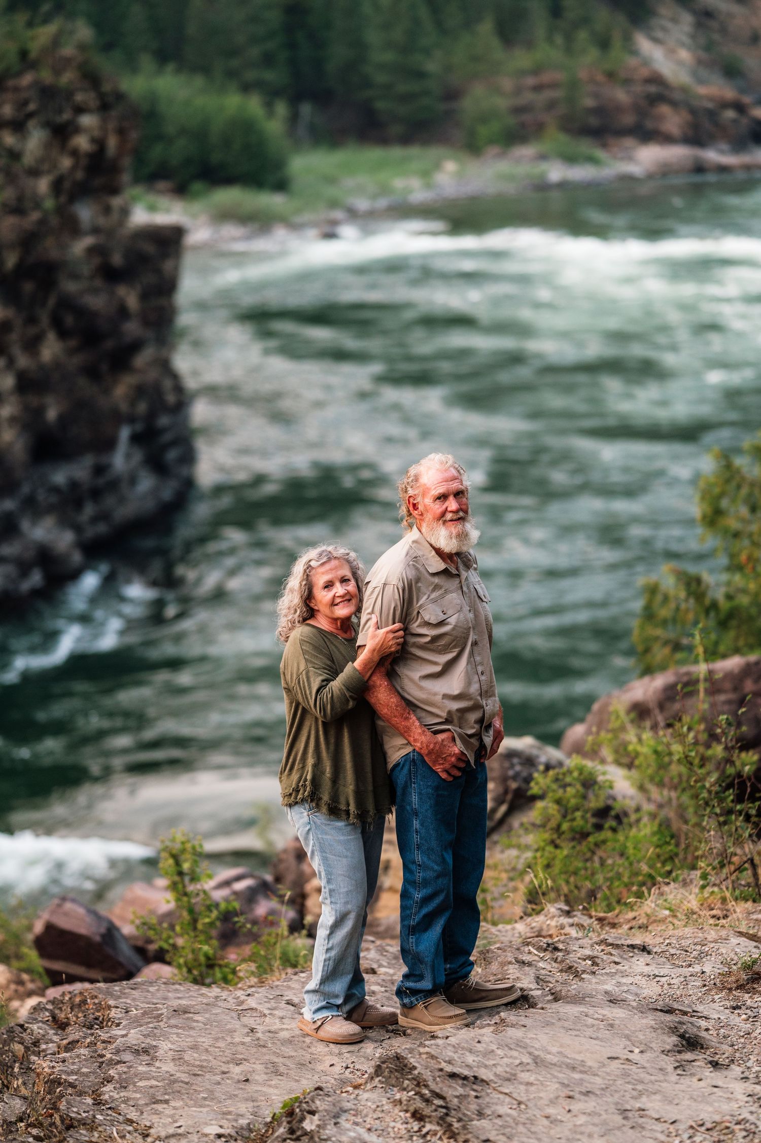 Senior couple standing on rocky overlook above a rushing river, woman embracing man from behind.