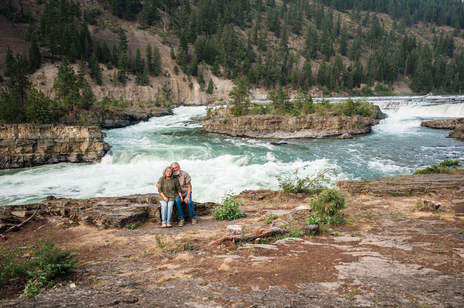 Elderly couple sitting close together on rocks overlooking a powerful waterfall in a scenic canyon.