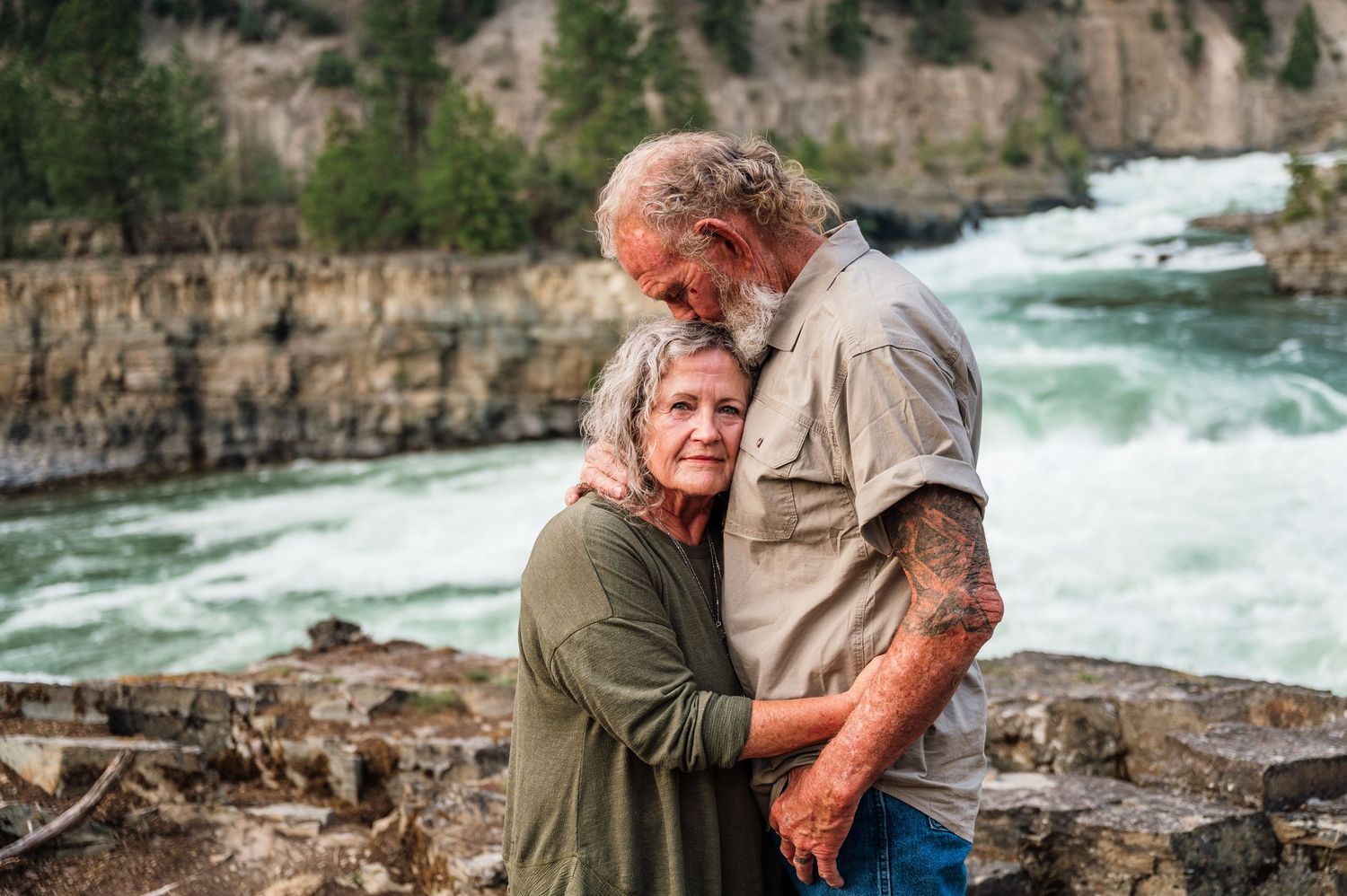 Elderly couple embracing romantically near a rushing river waterfall, man with tattoos holding woman in green shirt.