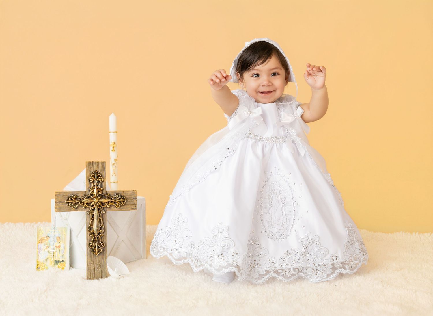 Baby girl in white lace christening gown and bonnet smiling beside a golden cross and candle.