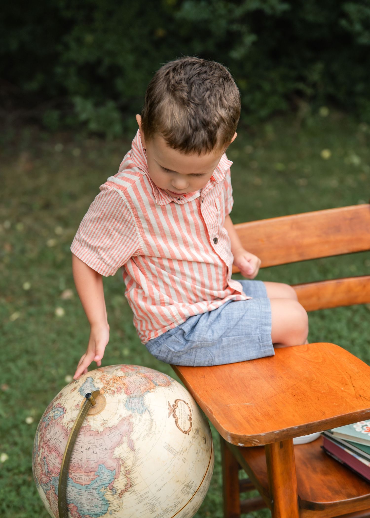 Excited young boy jumping in air outdoors, wearing blue shirt and khaki shorts, with globe and wooden chair on green lawn.
