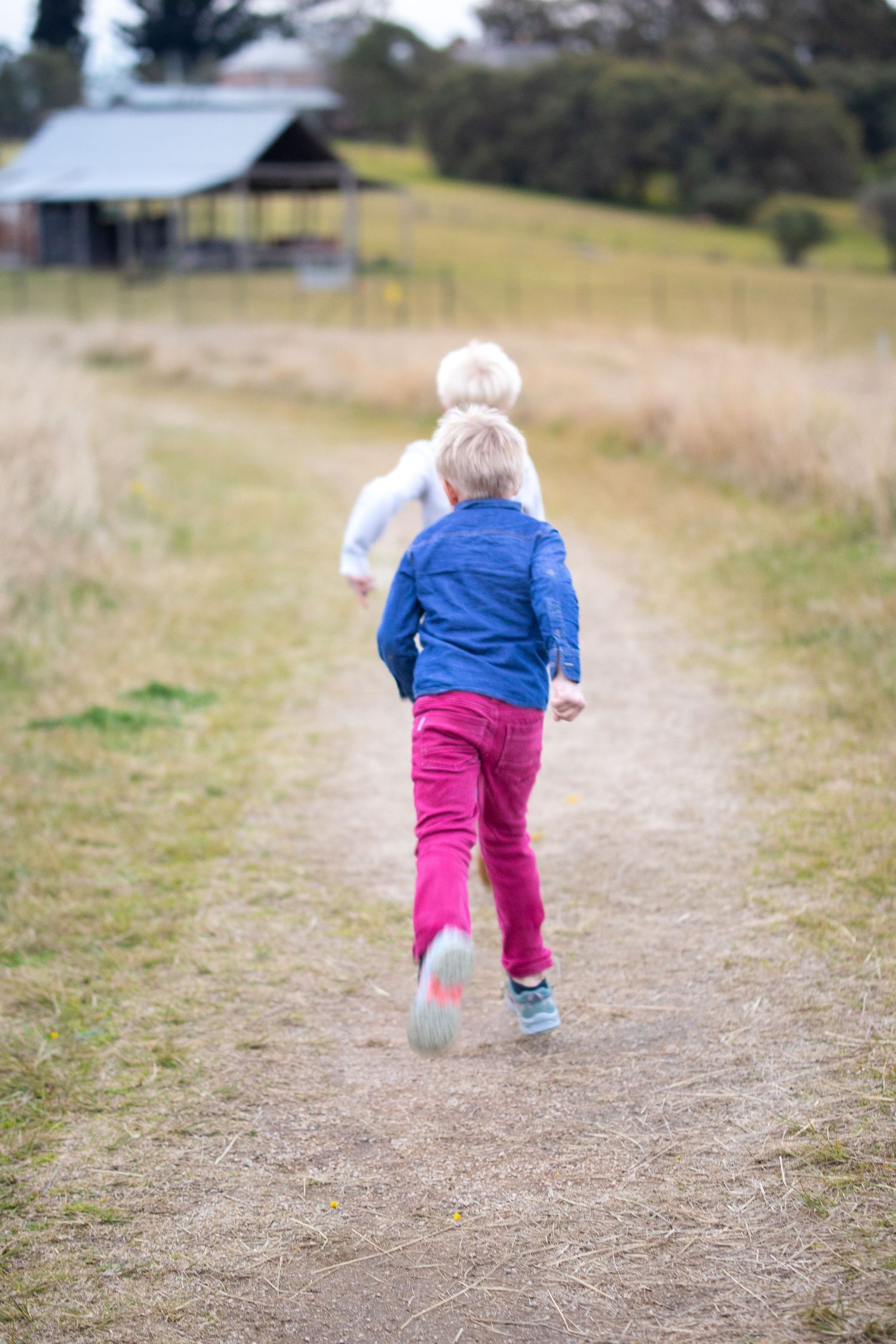 Two blonde children running down a rural dirt path surrounded by dry grass fields in Rouse Hill with a farm building in the background.