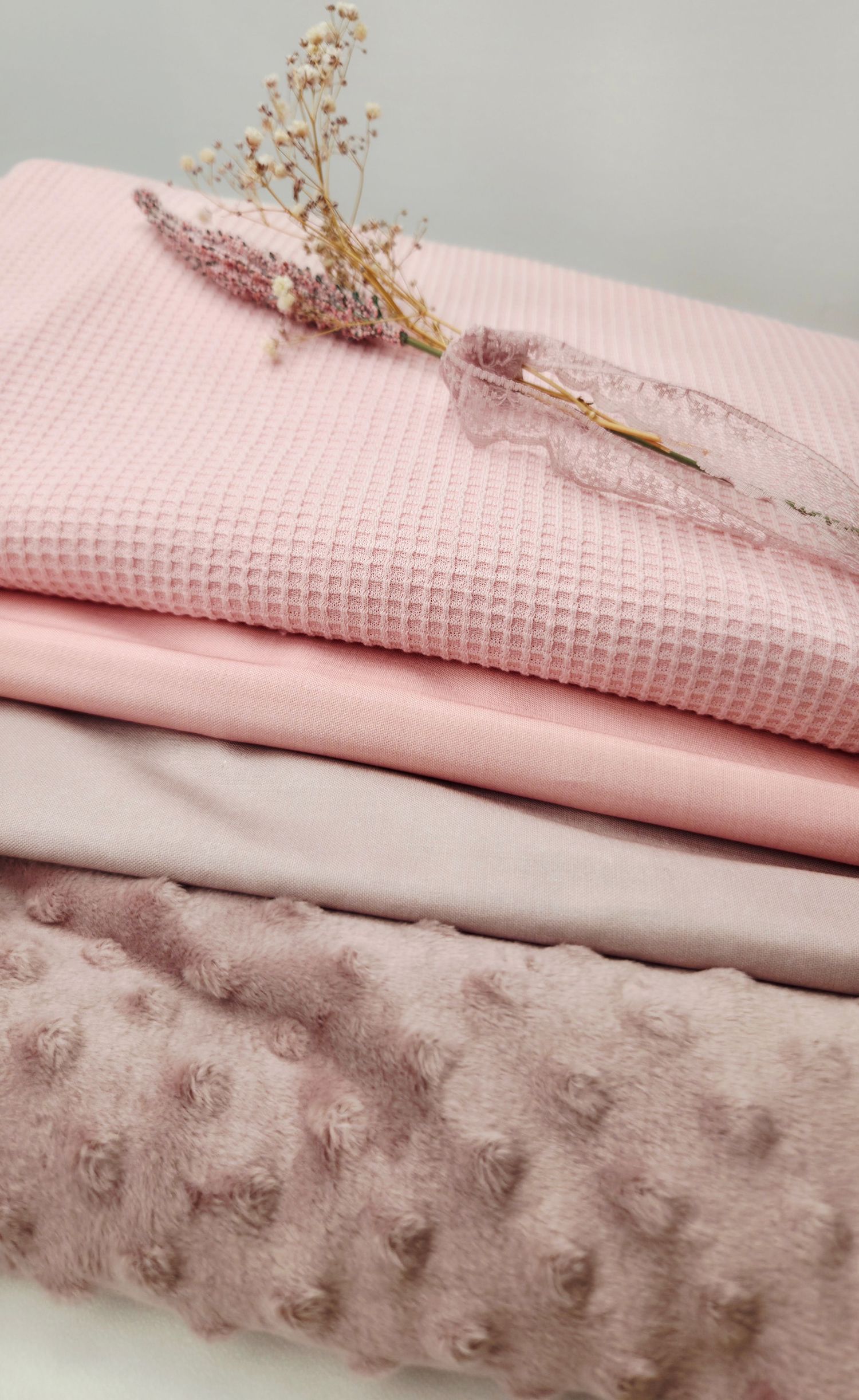 Stack of pink fabrics including waffle knit, smooth cotton, and minky dot textures, decorated with dried flowers.