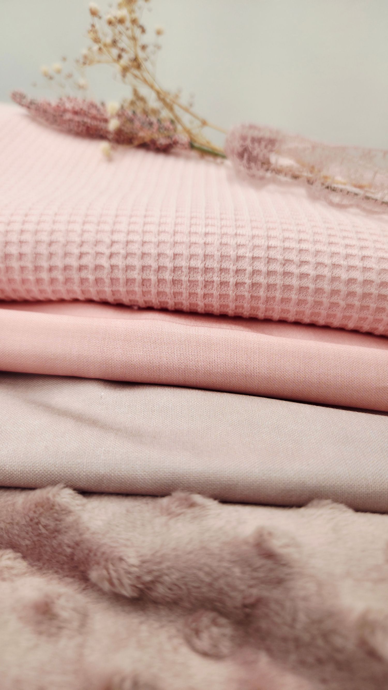 Stack of pink fabric swatches in waffle, jersey, and minky textures with dried flowers on white background.