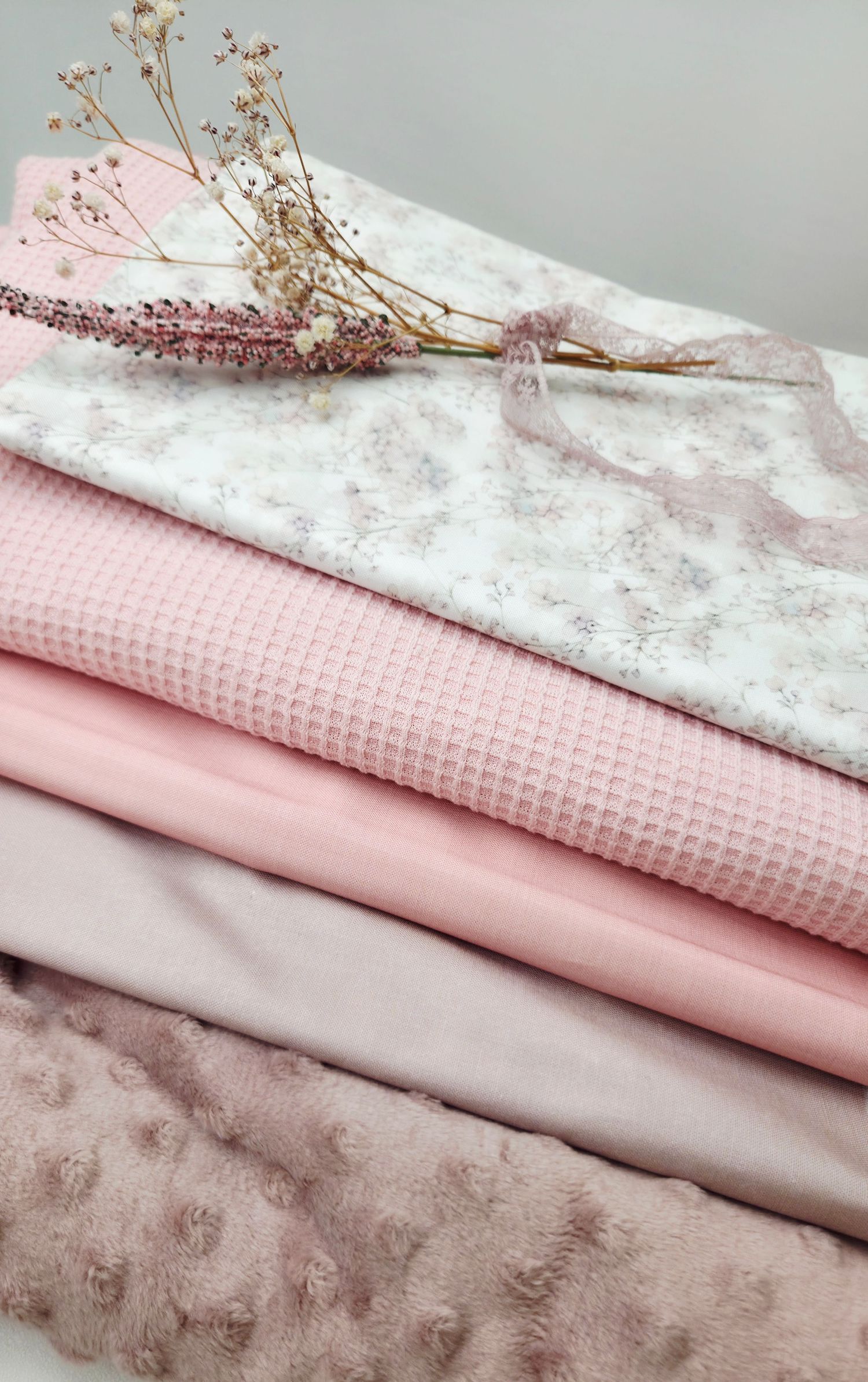 Stack of pink fabrics in various textures including waffle knit, minky, and floral print, styled with dried flowers.