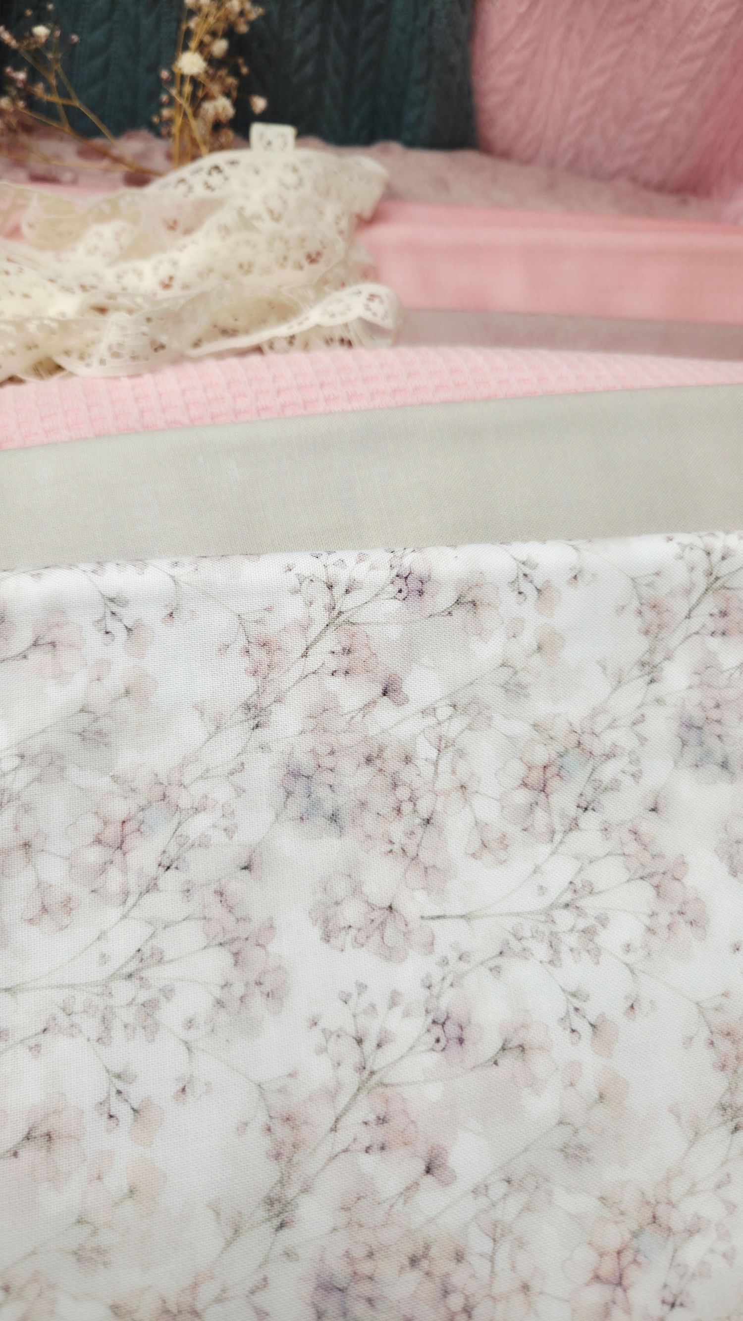 Delicate floral print fabric with soft pink cherry blossom pattern, displayed near lace trim and pink textiles.
