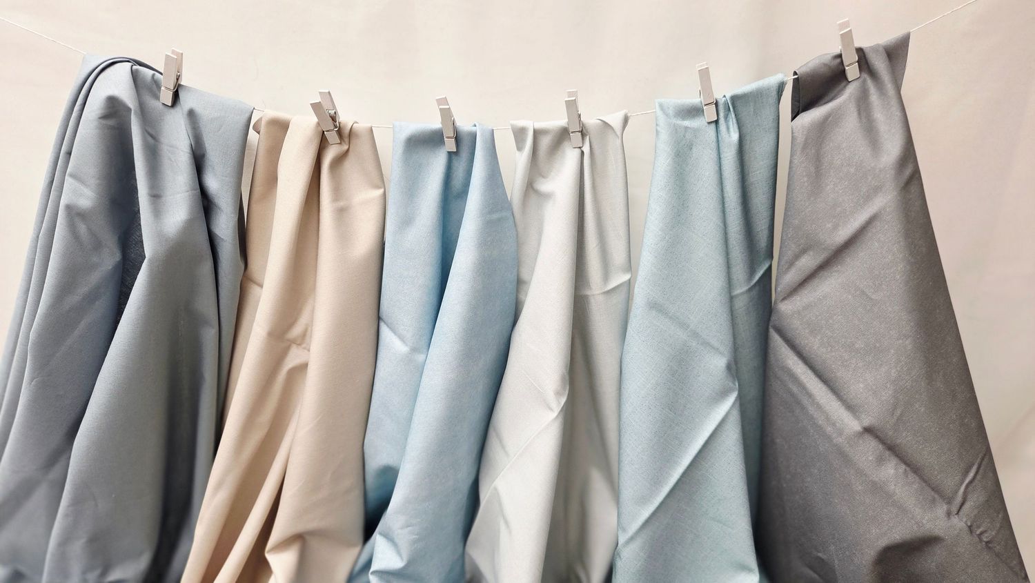 Collection of pastel fabric swatches in blue, beige and white tones hanging on clips against wall.